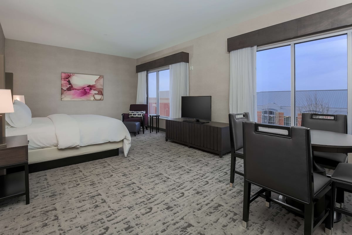 A hotel room with a bed, a desk, a TV, and large windows.