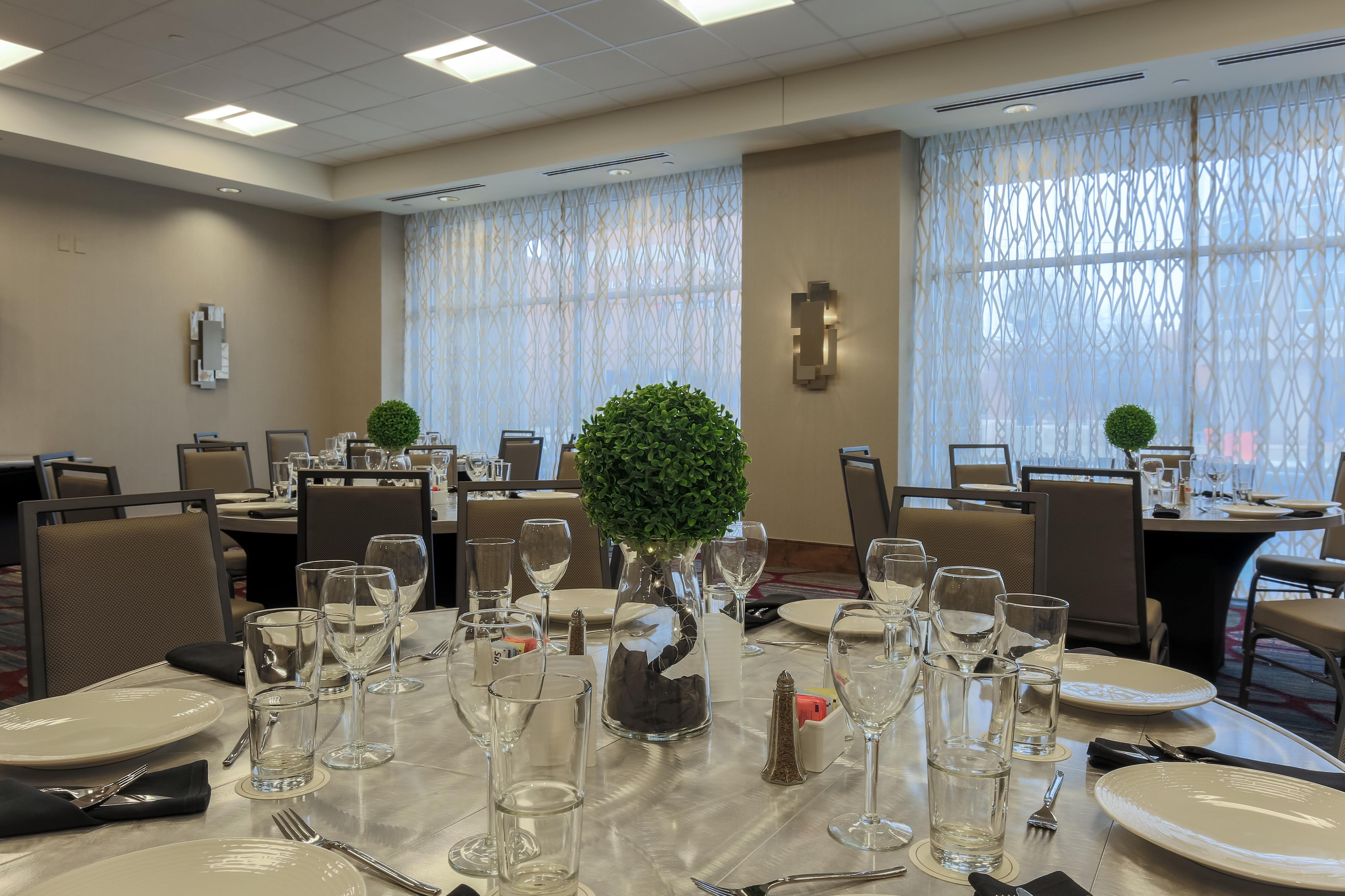 Un comedor con mesas dispuestas para una comida, con cristalería y platos transparentes, jarrones decorativos y un fondo de ventana con diseño.