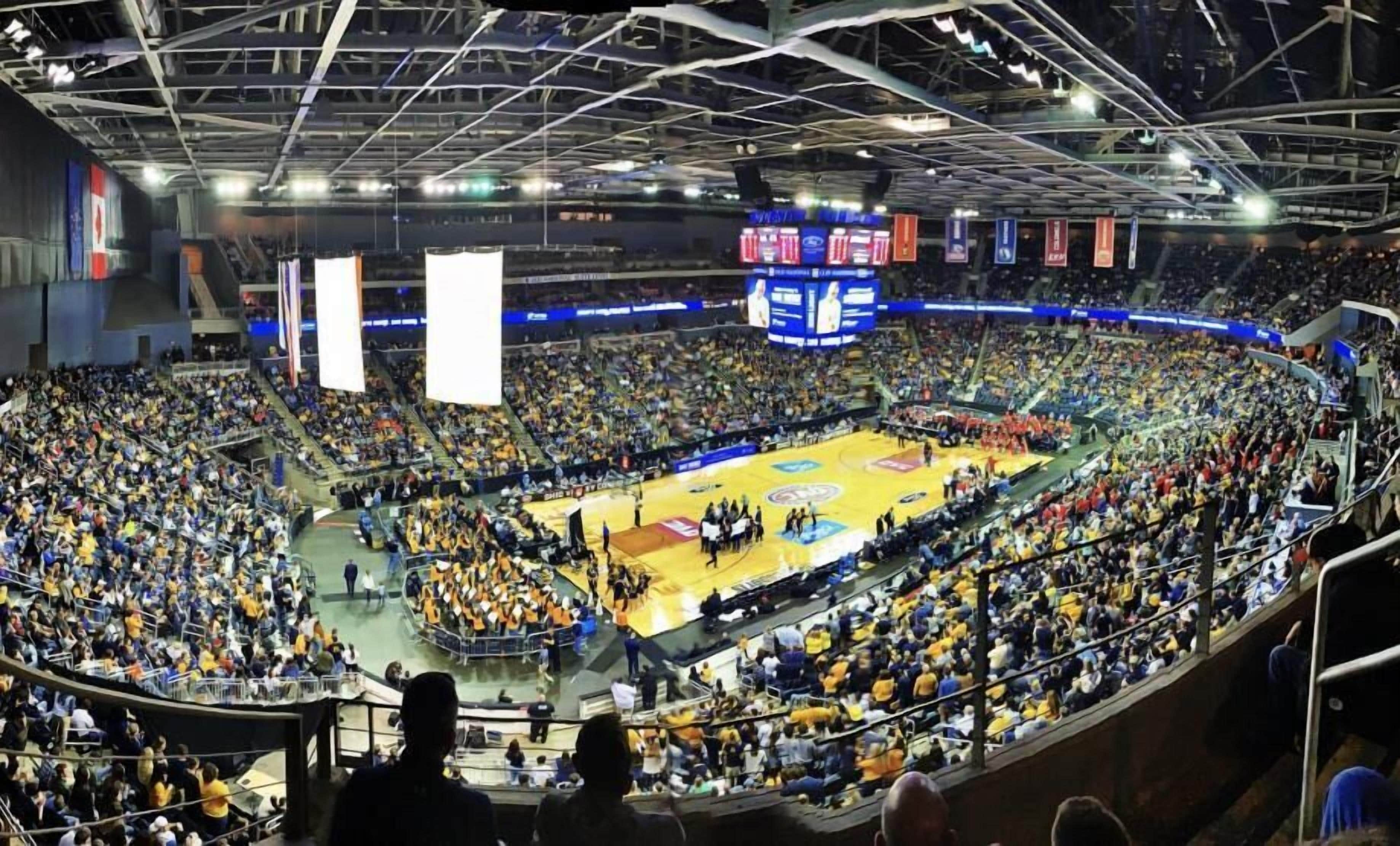 A large indoor stadium with multiple levels of seating, a central basketball court, and a scoreboard.