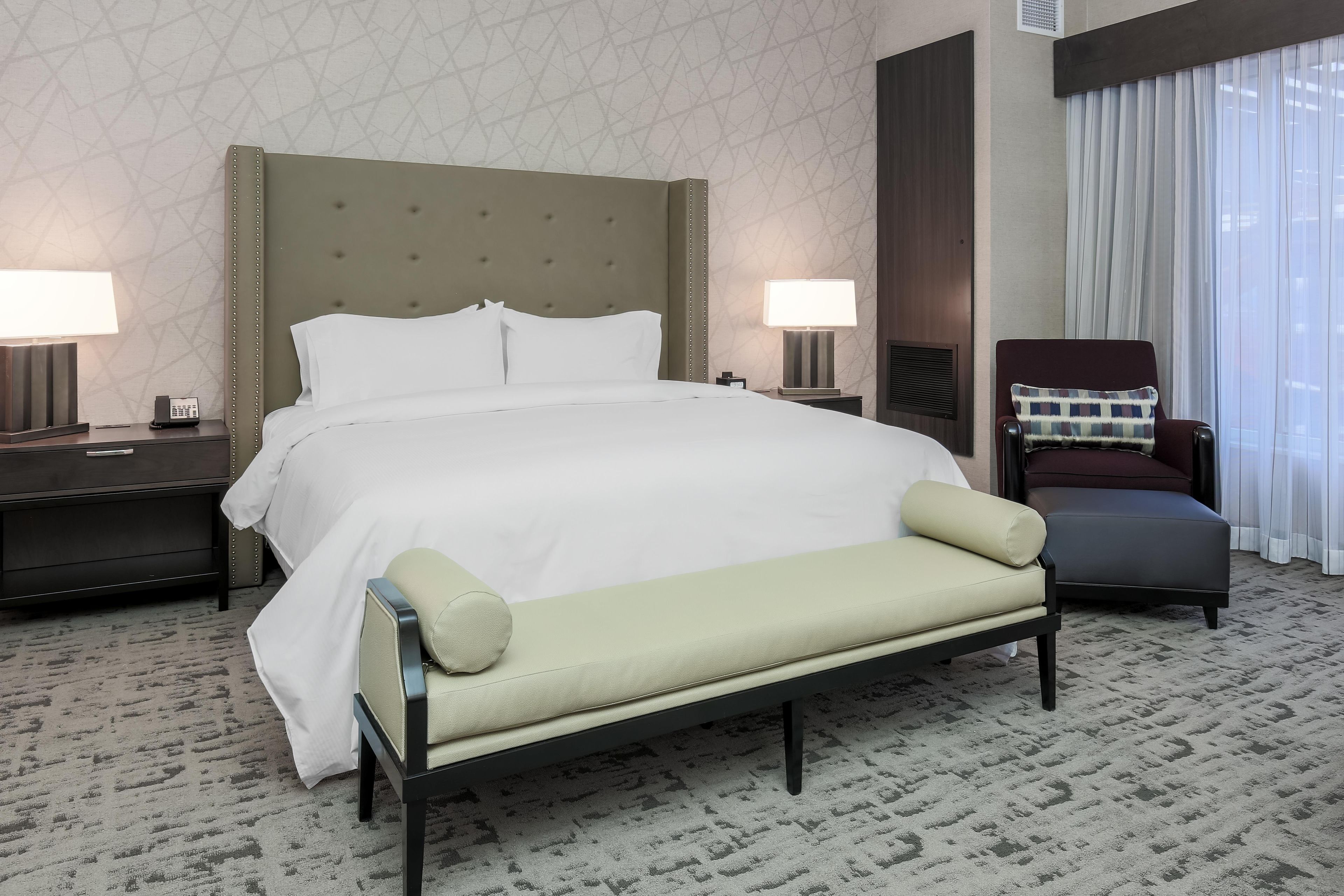 Une chambre d’hôtel avec un grand lit, deux tables de chevet avec des lampes, un banc, une chaise et une fenêtre avec des rideaux.