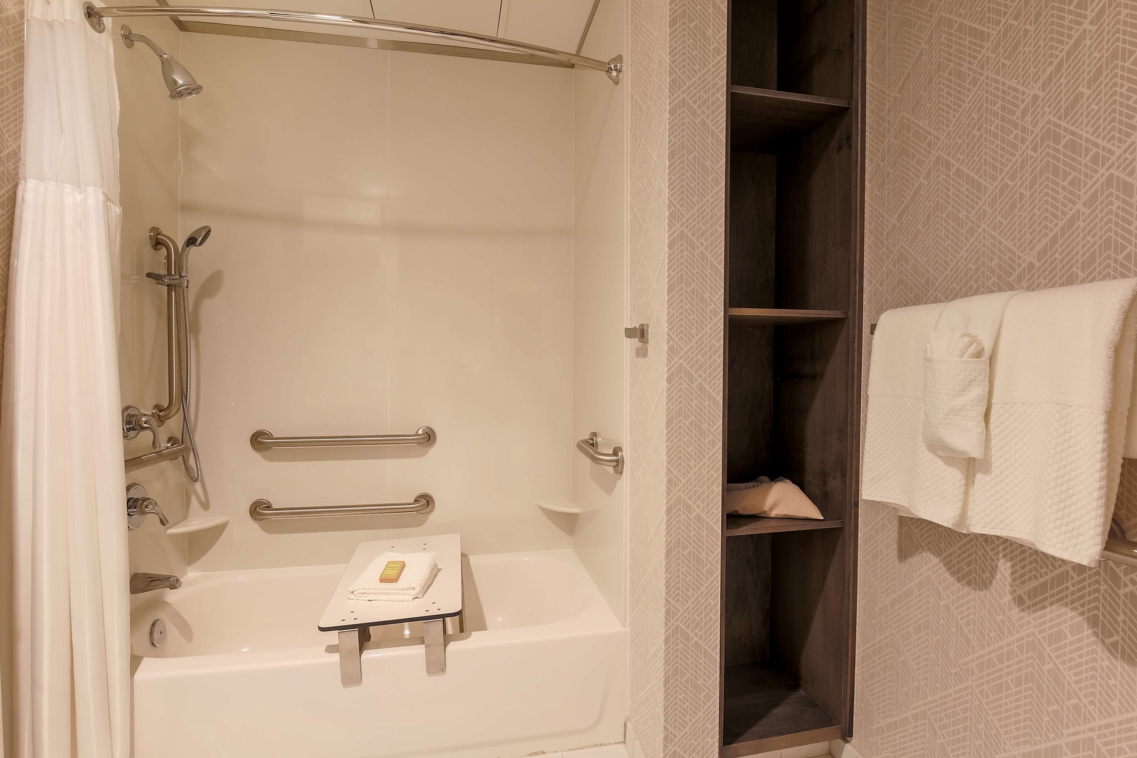 A bathroom with a bathtub, shower, and handrails. There is a towel rack with towels and a small shelf with toiletries.