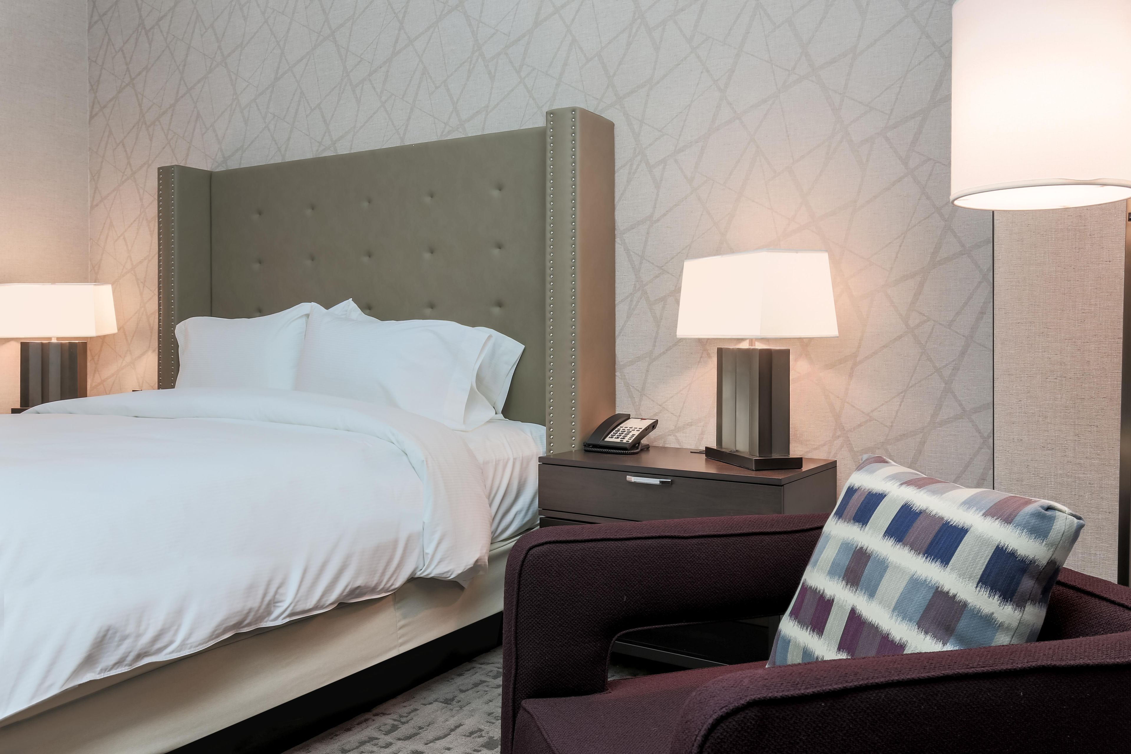A hotel room with a bed, two bedside lamps, a telephone, and a chair with a patterned cushion.
