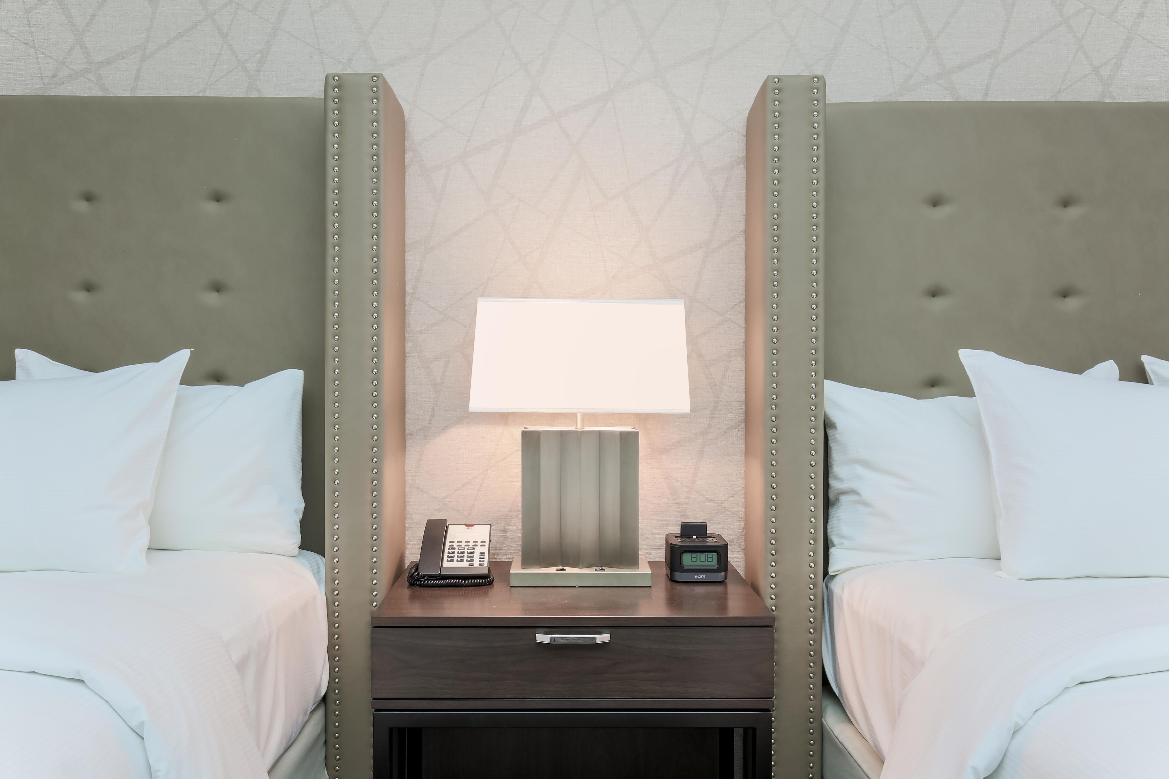 Habitación de hotel con dos camas, una mesita de noche con lámpara, teléfono y reloj digital.
