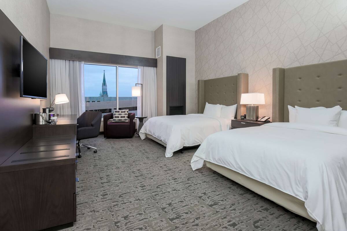 A hotel room with two beds, a desk, a TV, and a view of a church spire.