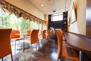 Un espace repas moderne avec des chaises et des tables orange, une grande fenêtre avec des rideaux et un téléviseur fixé au mur.