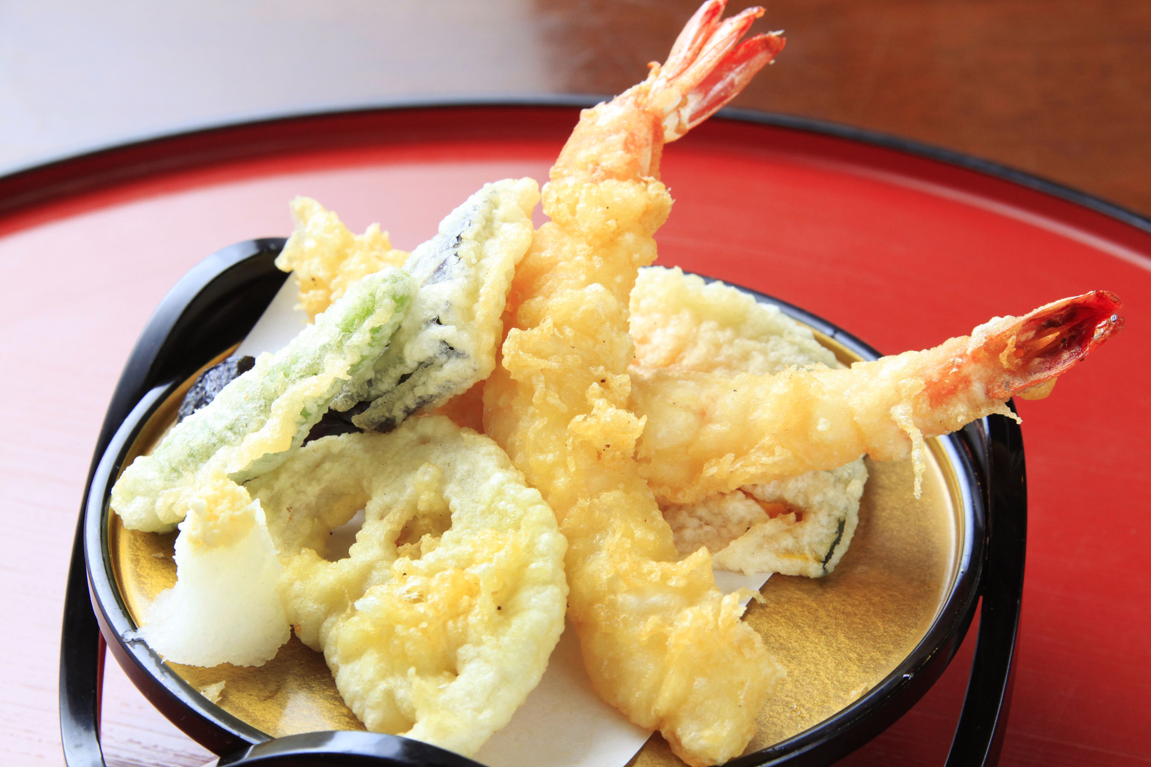 A bowl of tempura shrimp and vegetables on a black plate with a red background.