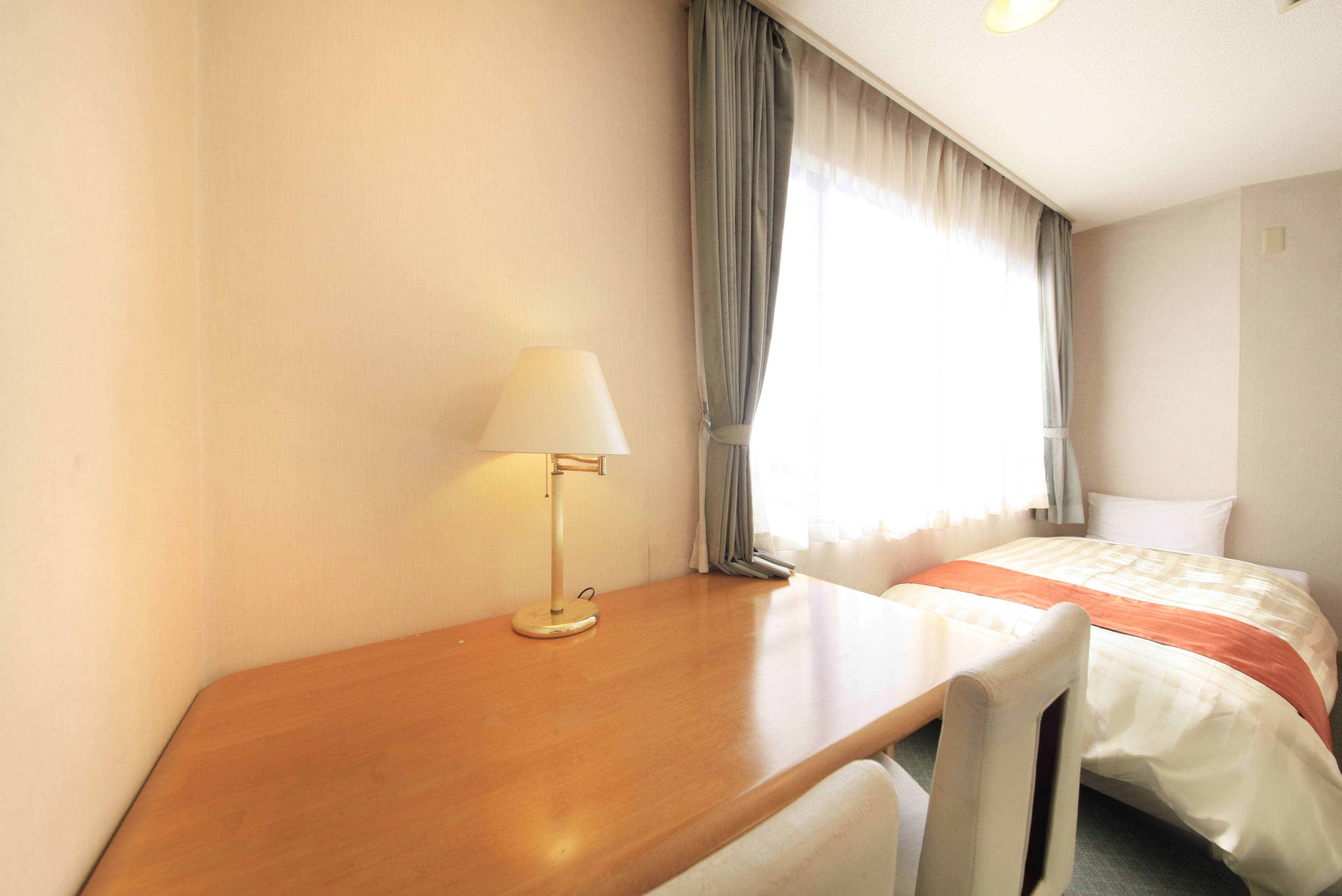Habitación de hotel con escritorio de madera, lámpara, cama con colcha blanca y naranja, y ventana con cortinas.