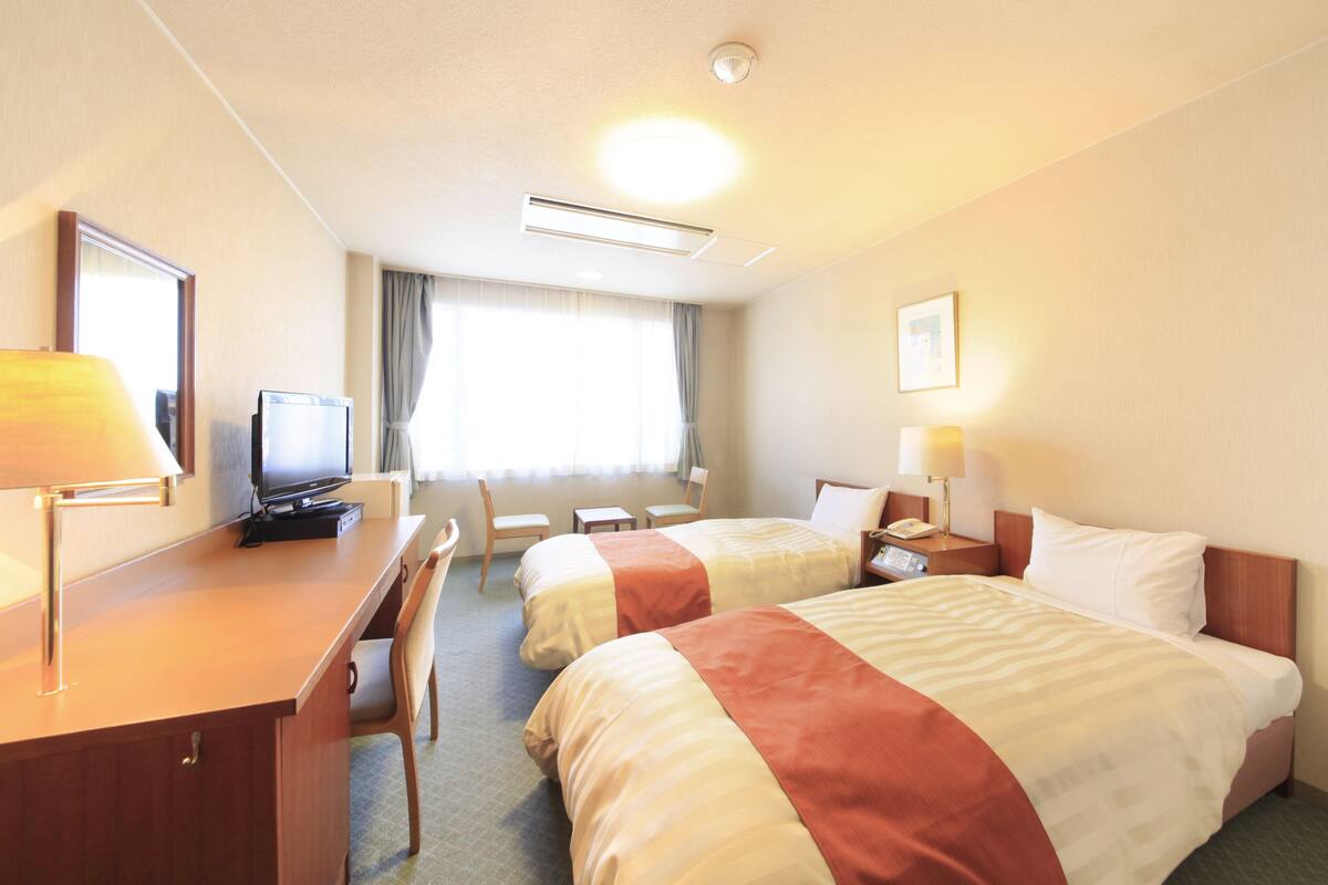 A hotel room with two beds, a desk with a computer, a chair, a TV, and a window with curtains.