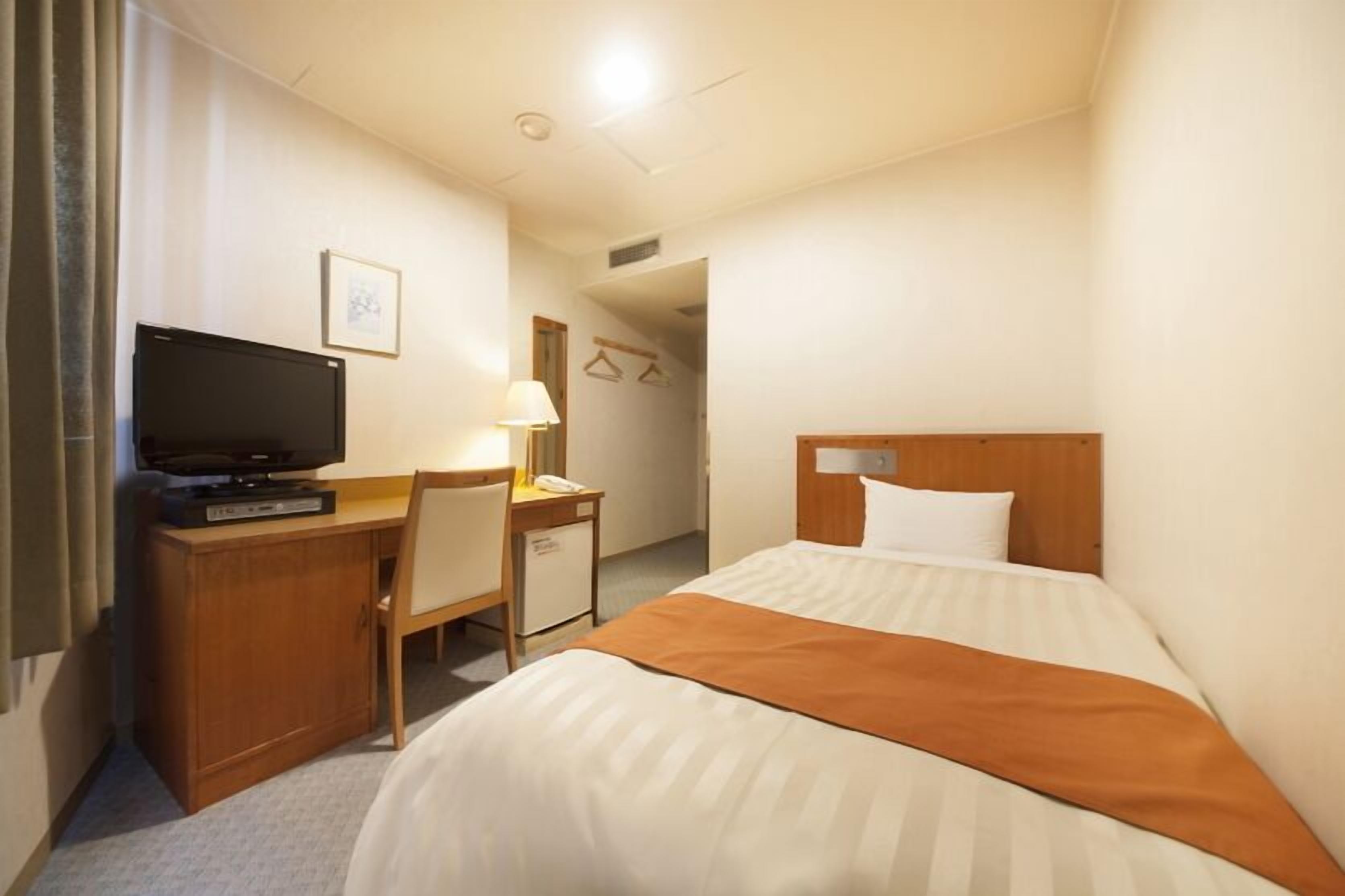 A hotel room with a bed, a desk with a television, a lamp, and a wardrobe.