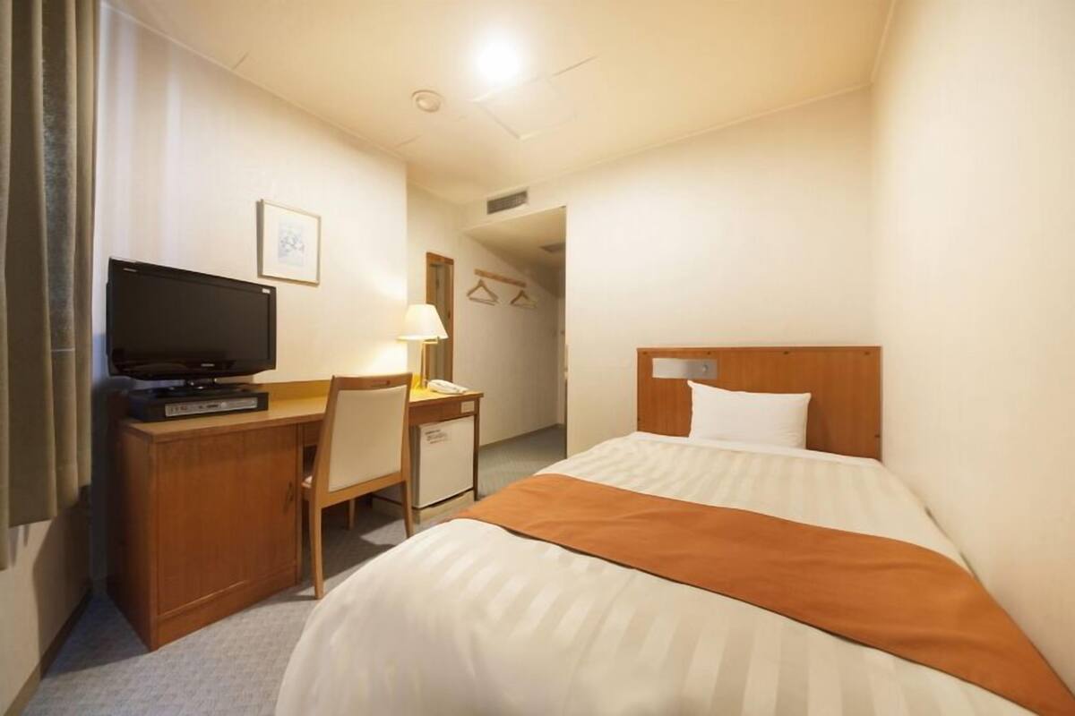 A hotel room with a bed, a desk with a television, a lamp, and a wardrobe.