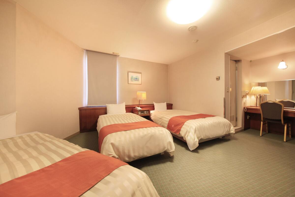 A hotel room with three beds, a desk, a chair, and a lamp.
