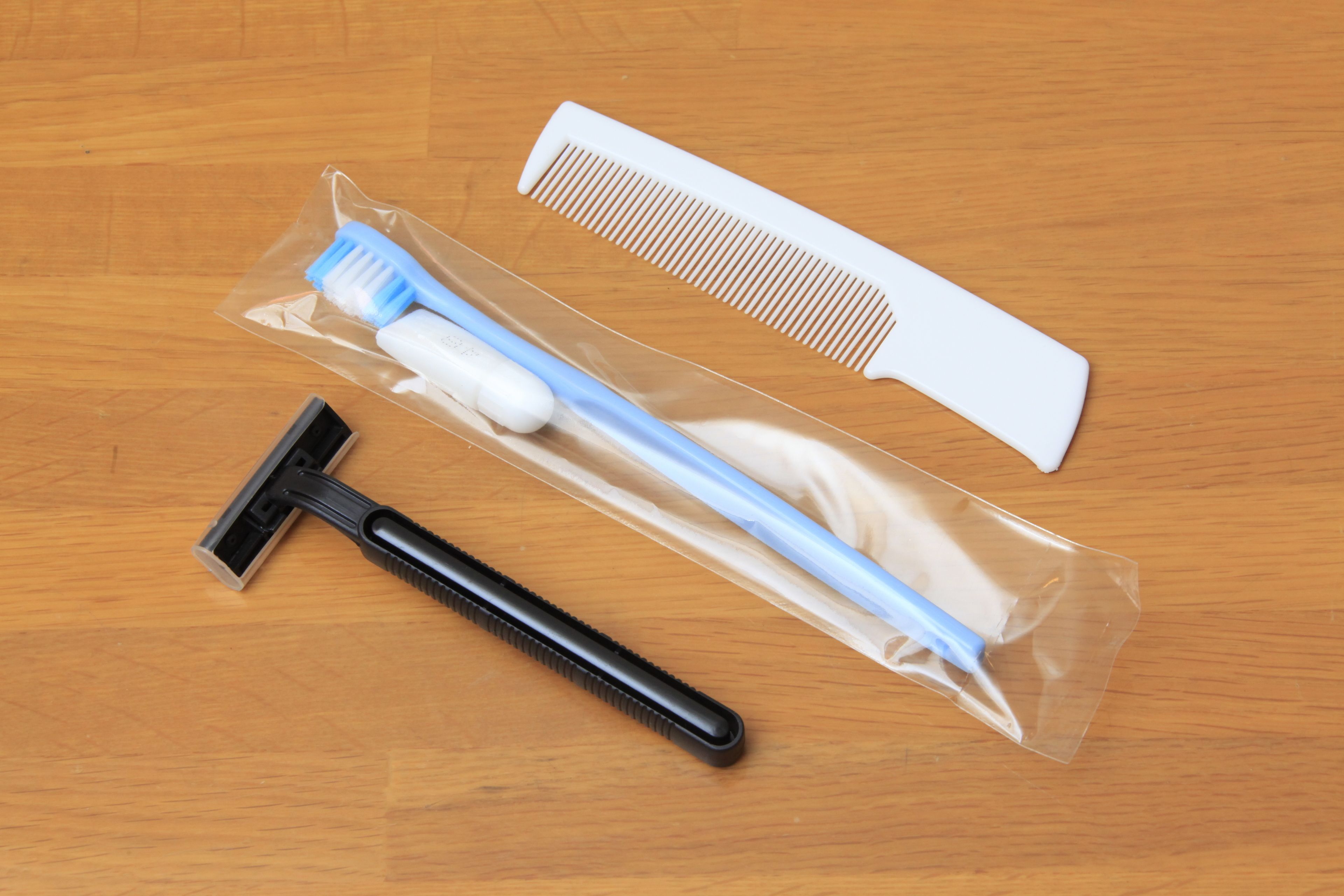 Embalagem contendo uma escova de dentes preta e azul, um pente branco e uma capa para escova de dentes preta e azul sobre uma superfície de madeira.