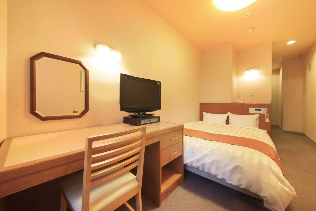 A hotel room with a wooden desk, a television, a bed with white bedding, and a mirror on the wall.