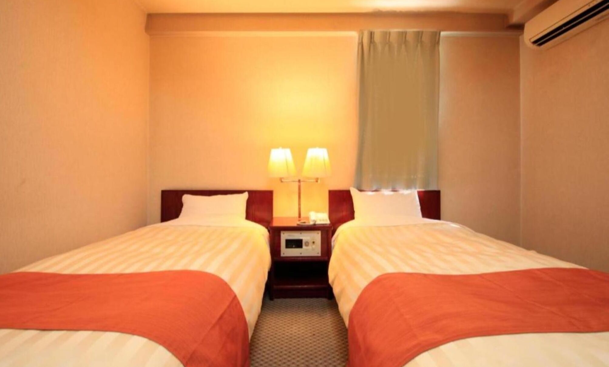 A hotel room with two beds, a bedside table with lamps, and a window with a curtain.