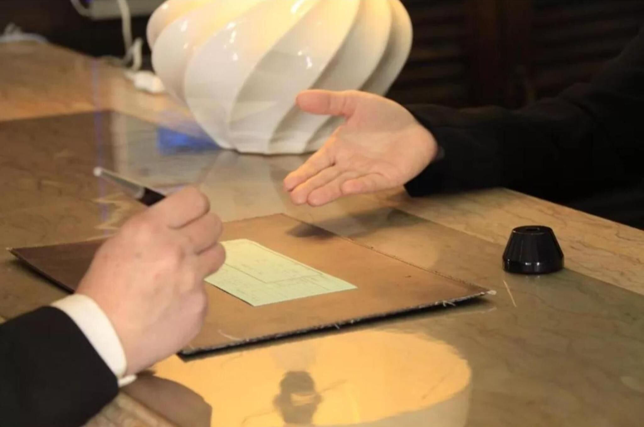 A person signing a document on a table with a laptop and a small black object.
