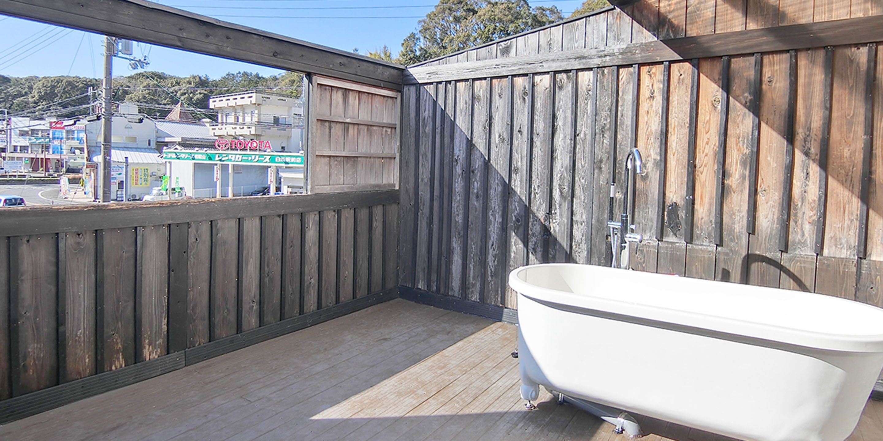Una terraza de madera con una bañera blanca, paredes de madera y vistas a una zona comercial con un letrero de 'TOYOTA'.