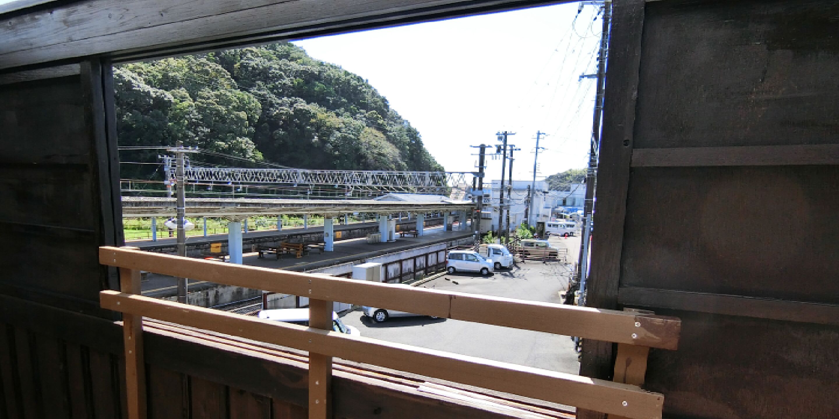 Vista desde el interior de un edificio, mostrando una estación de tren con vías, una camioneta blanca y un paisaje montañoso al fondo.