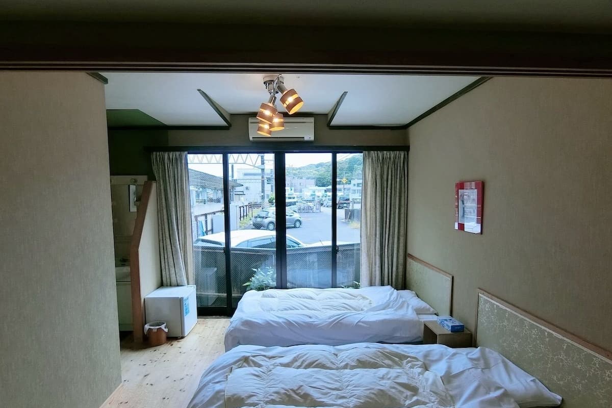 Een hotelkamer met twee bedden, een groot raam met uitzicht op de straat, een plafondlamp en een klein tafeltje met nachtkastje.
