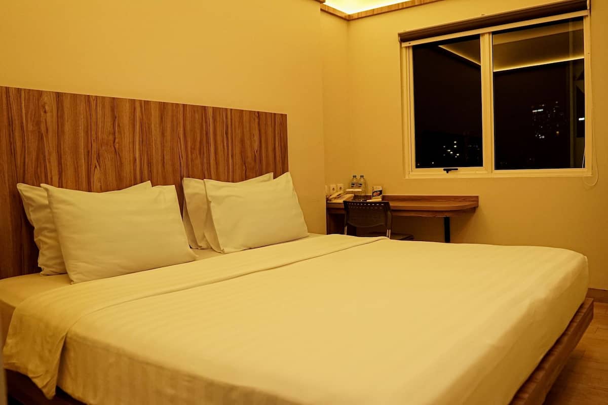A neatly made bed with white linens and pillows, a wooden headboard, a small desk with a chair, a window with a view of the night cityscape, and a wooden floor.