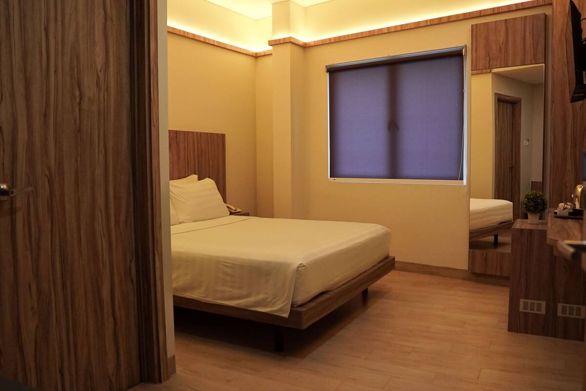 A bedroom with a bed, a bedside table, a mirror, and a window with blinds.