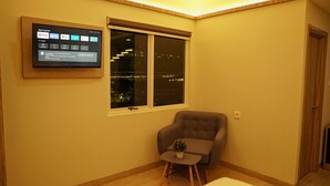 A room with a flat-screen TV mounted on the wall displaying apps and games. There is a window with a view of the city at night. A small table with a plant is placed in front of a grey armchair. - Hotel Cikini (Jakarta)