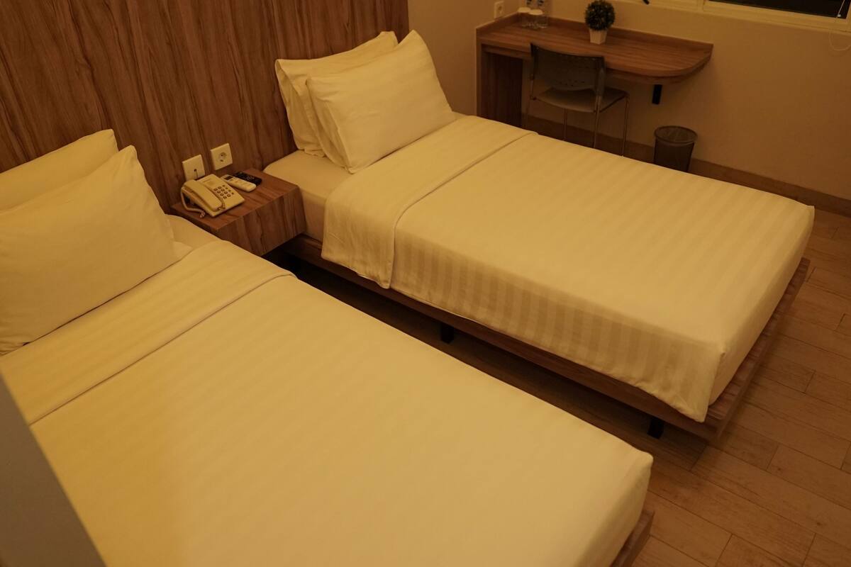 A hotel room with two single beds, a wooden headboard, a bedside table with a phone and remote control, a small desk with a chair, and a window with a plant on the sill.