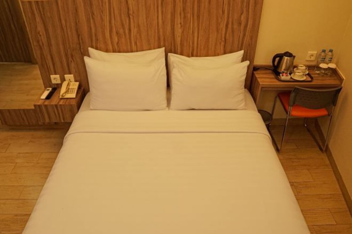 A hotel room with a bed, a wooden headboard, a bedside table with a phone, a kettle, and a tray with cups and bottles.