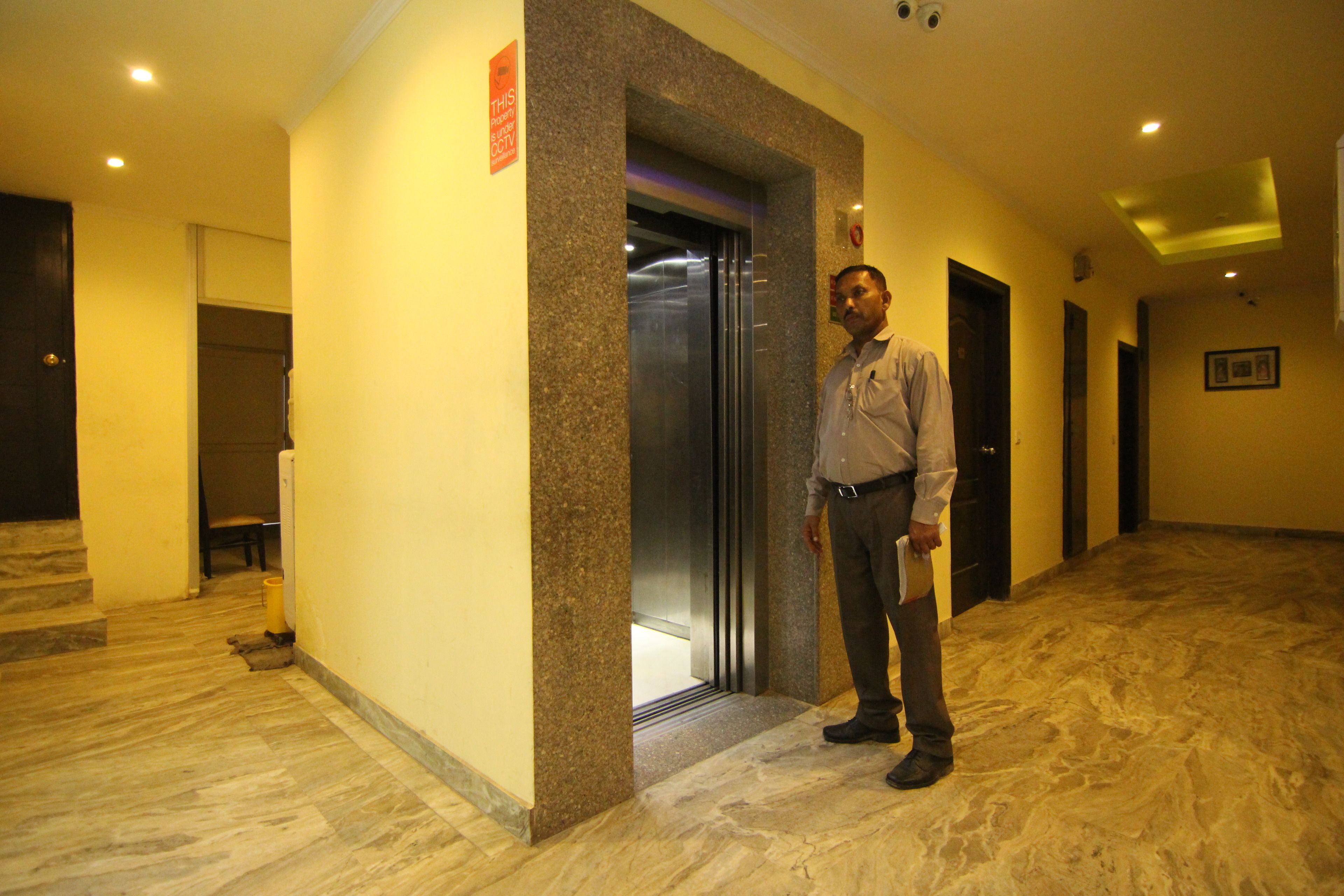 A man standing in a hallway with a marble floor, a door, and an elevator.