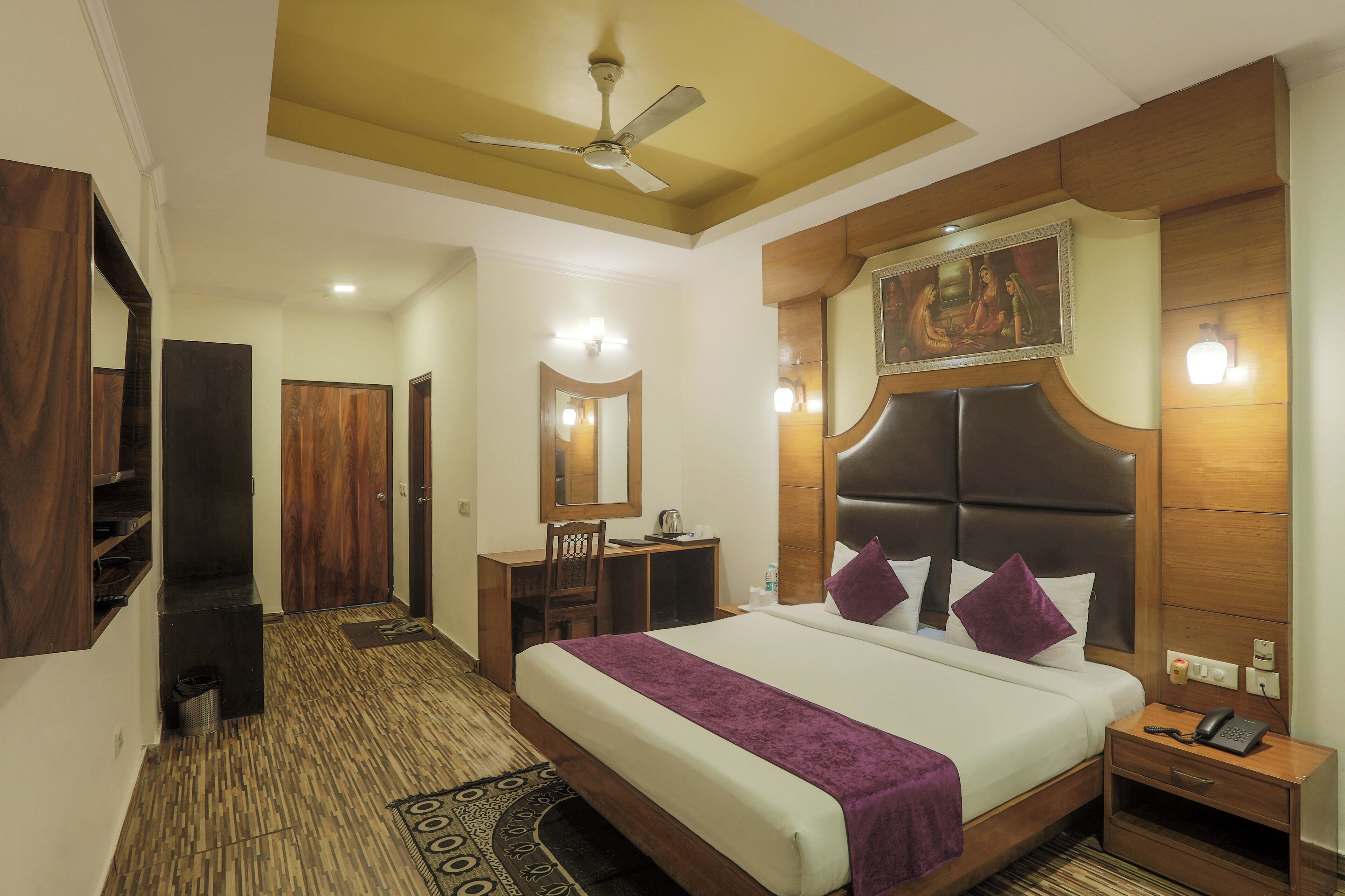 Une chambre d’hôtel avec un grand lit, un bureau, une chaise, une armoire, un ventilateur de plafond et un tableau au mur.