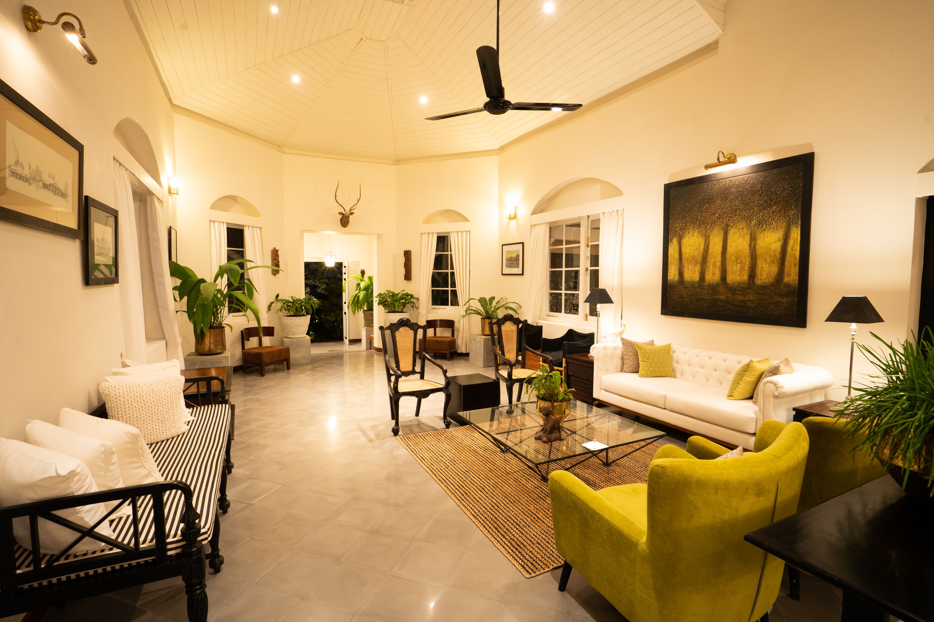 A spacious living room with a high ceiling, a ceiling fan, and a large painting on the wall. There are several chairs and a sofa arranged around a coffee table.