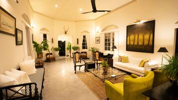 A spacious living room with a high ceiling, a ceiling fan, and a large painting on the wall. There are several chairs and a sofa arranged around a coffee table.