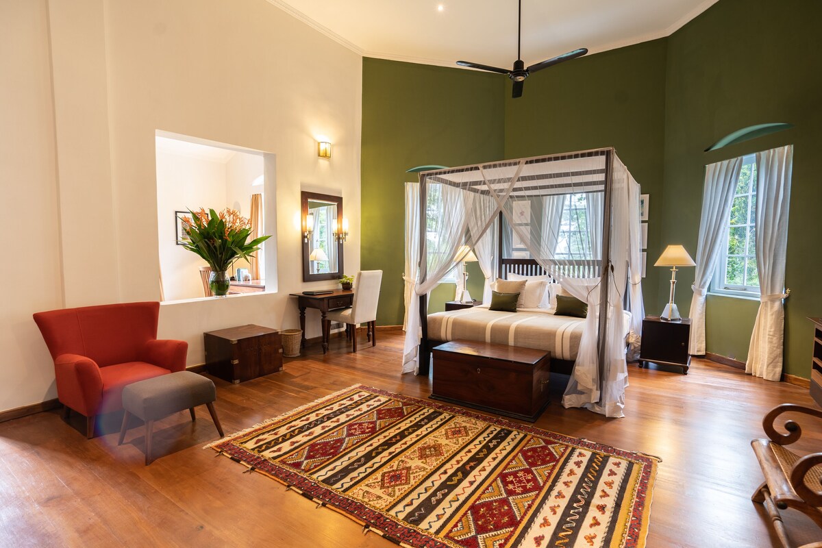 A spacious bedroom with a four-poster bed, a red armchair, a wooden bench, and a patterned rug.