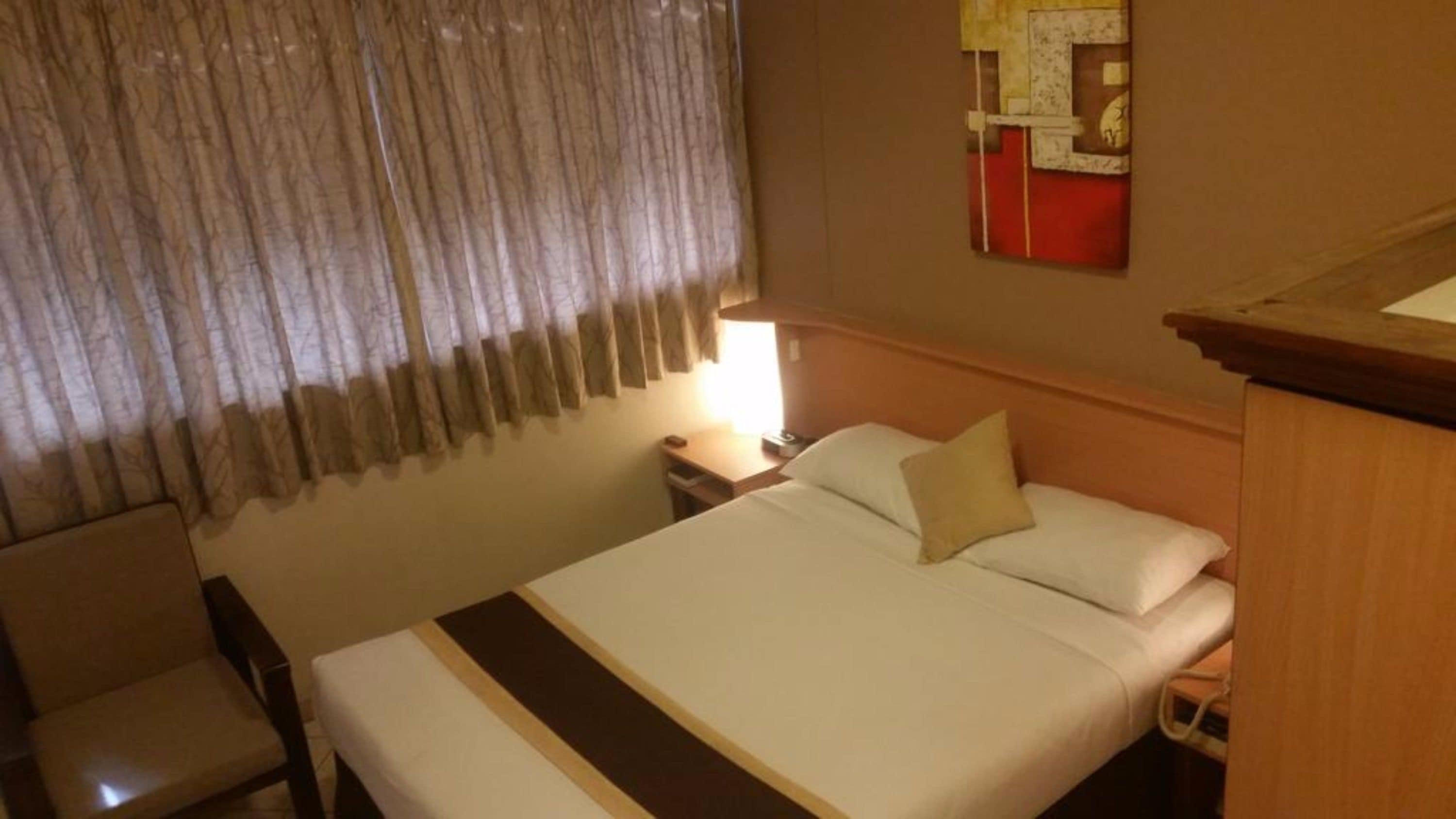 A hotel room with a bed, a nightstand, a lamp, a chair, and a window with curtains.