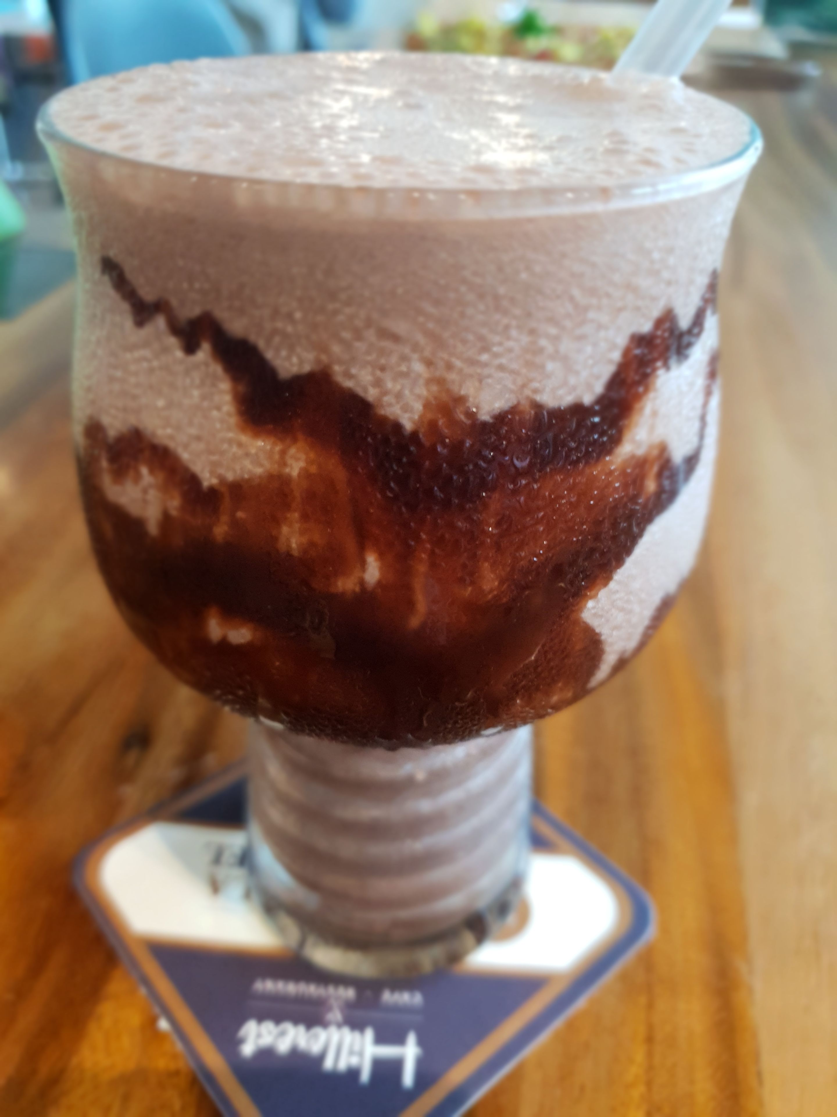 Une boisson au chocolat à plusieurs couches, servie dans un verre avec une paille, posée sur un dessous de verre orné d’un texte.