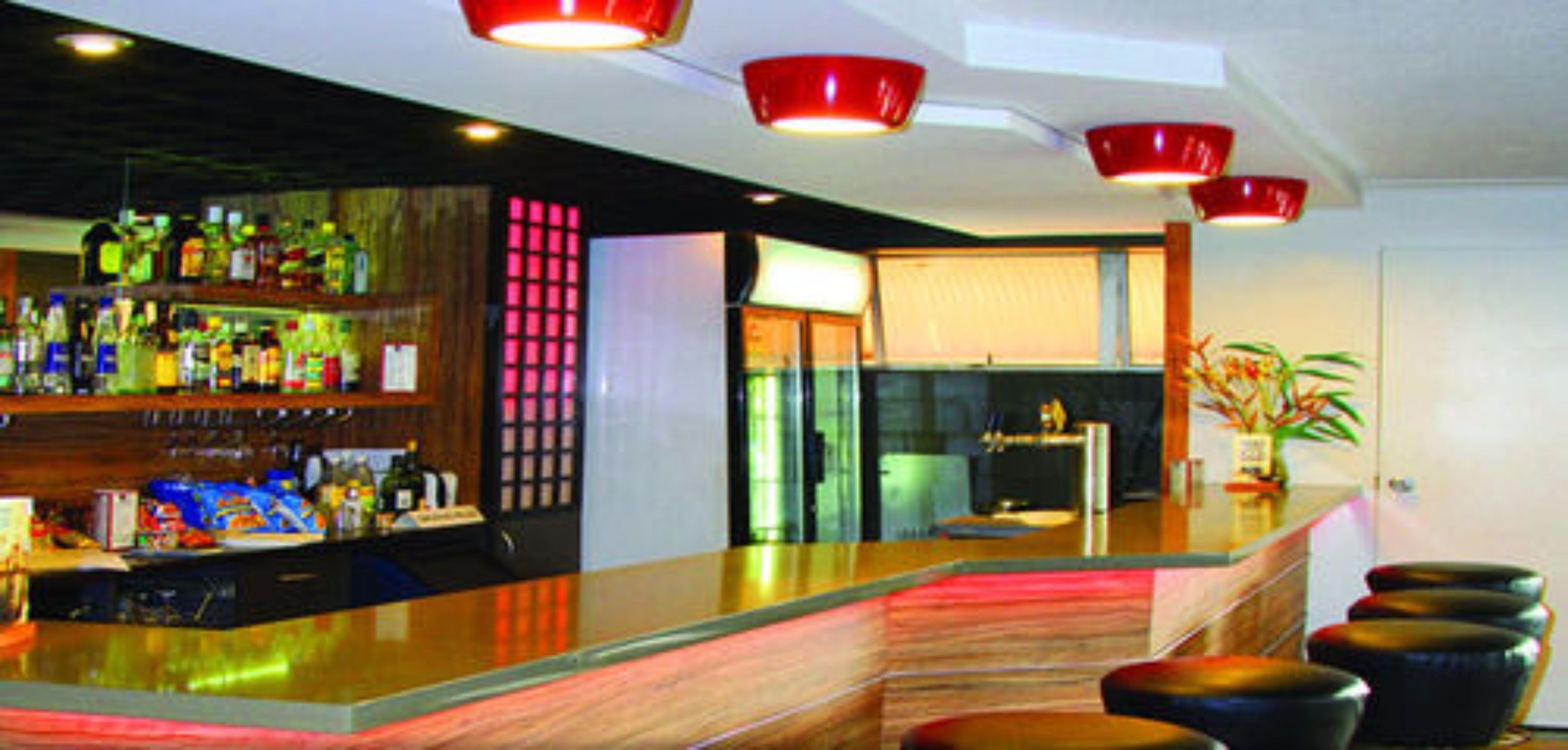 Zona de bar moderna con mostrador de madera, luces colgantes rojas y una variedad de botellas.