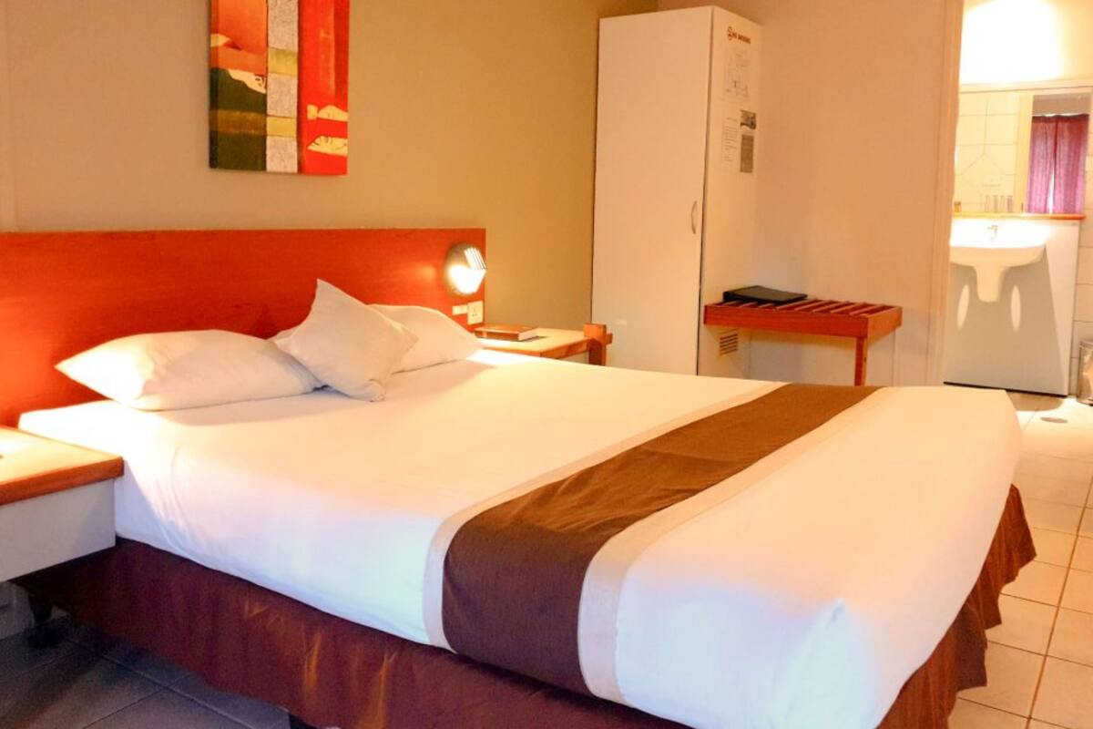 A hotel room with a large bed, a nightstand, a wardrobe, and a small table with a book.