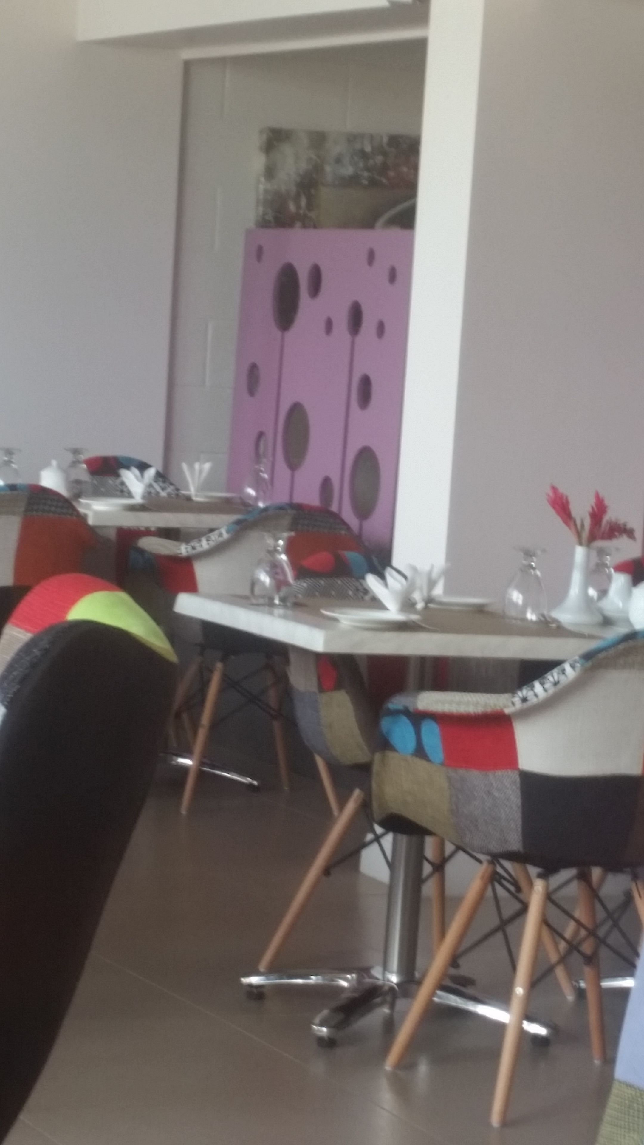 Un coin repas avec des chaises et des tables aux couleurs vives, un mur rose orné d’œuvres d’art abstraites et une porte visible au fond.