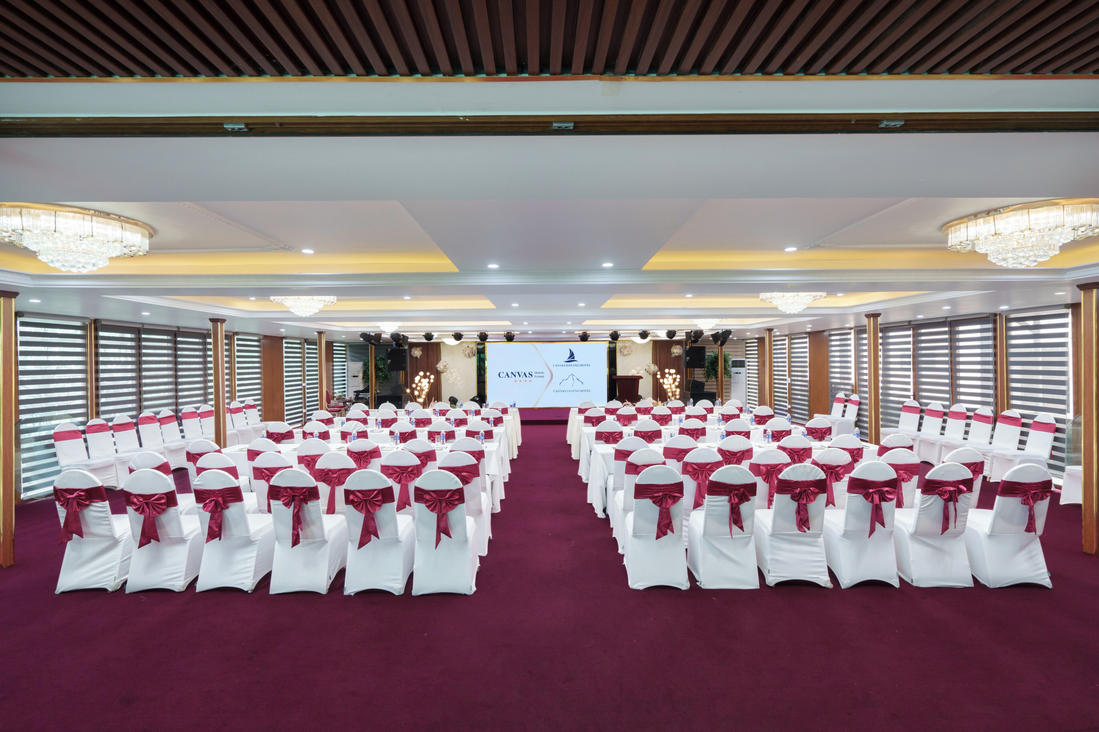 A conference room with rows of white chairs with red sashes, a large screen at the front, and chandeliers on the ceiling.