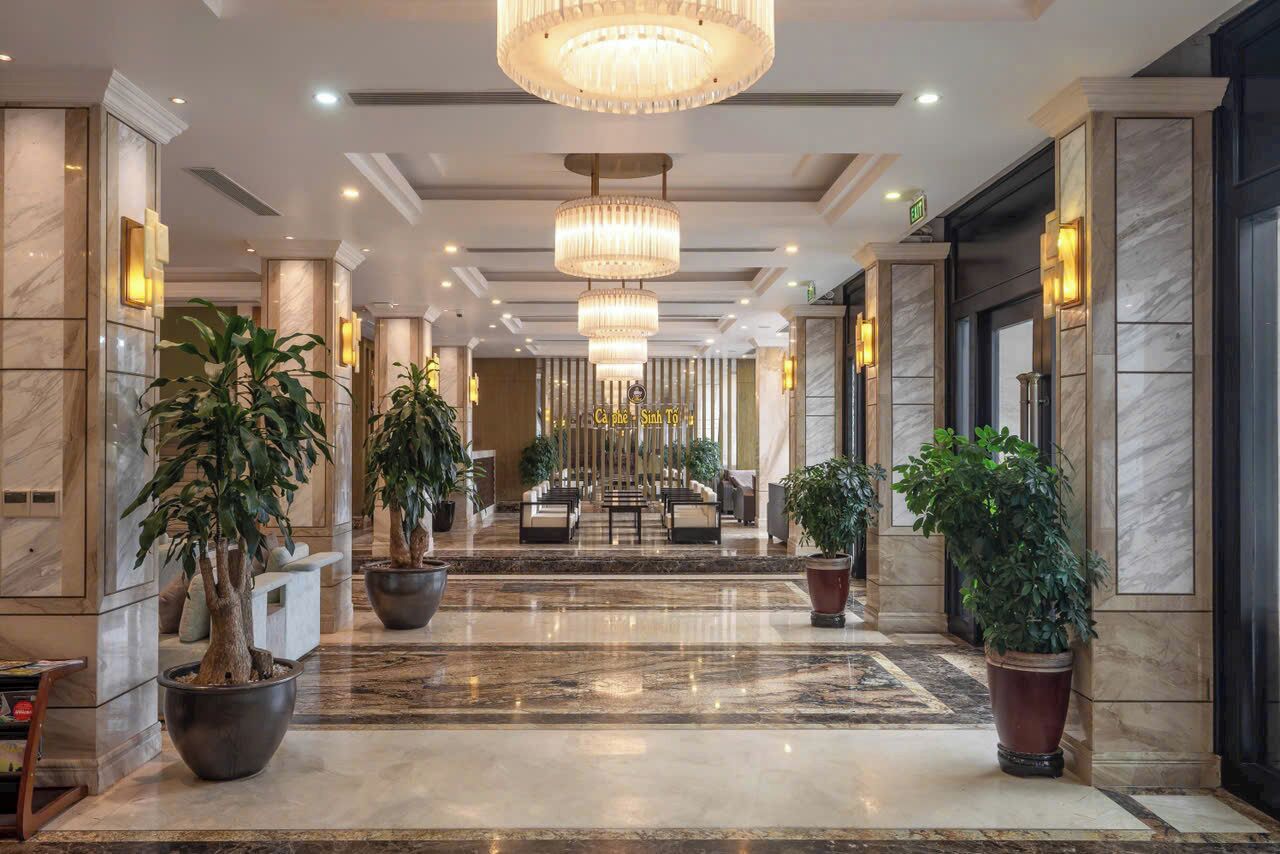 Un vestíbulo de hotel con suelo de mármol, plantas en macetas, un mostrador de recepción y una araña.