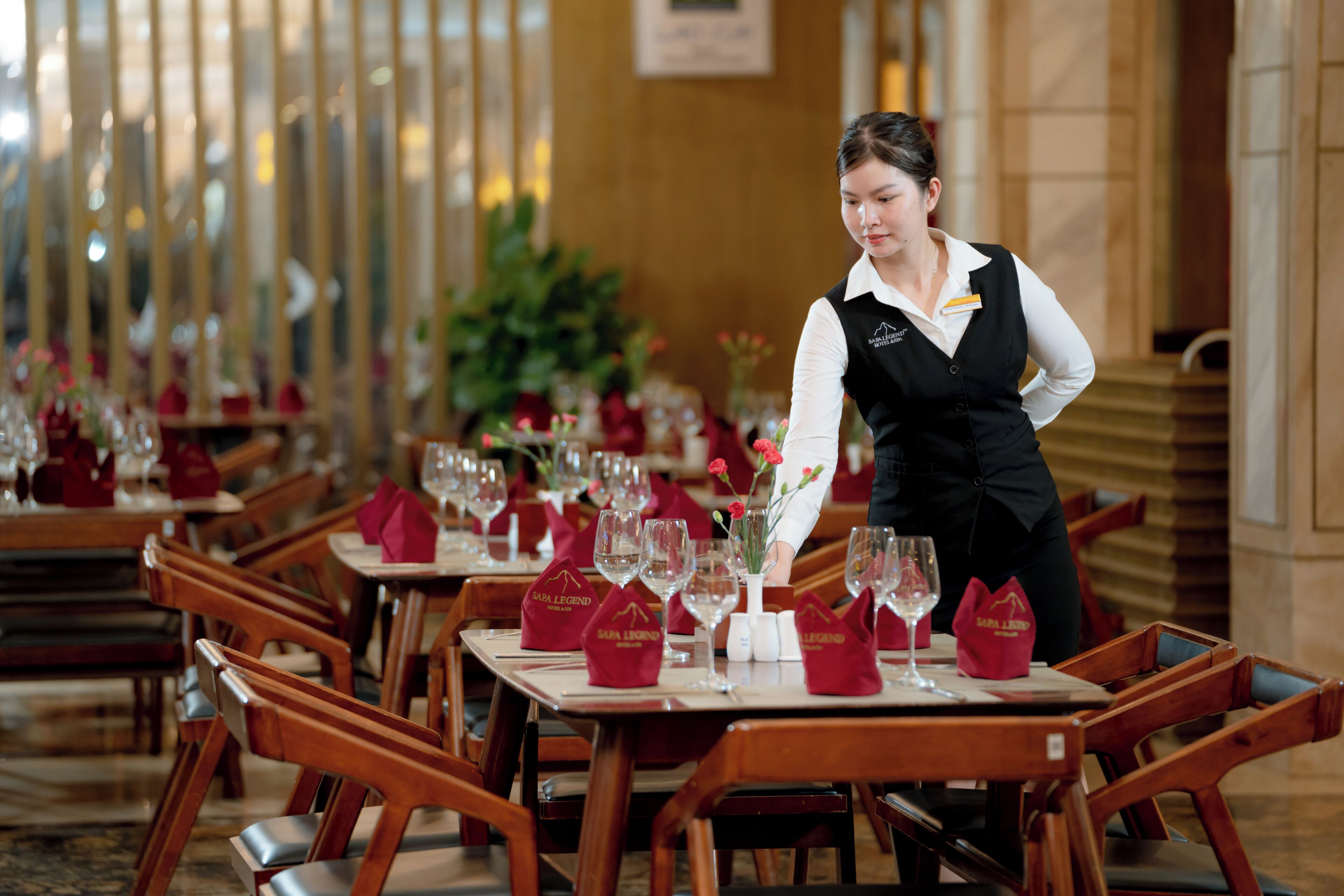 A hotel dining area with tables set for guests, a waiter attending to a table, and a decorative vase with flowers.