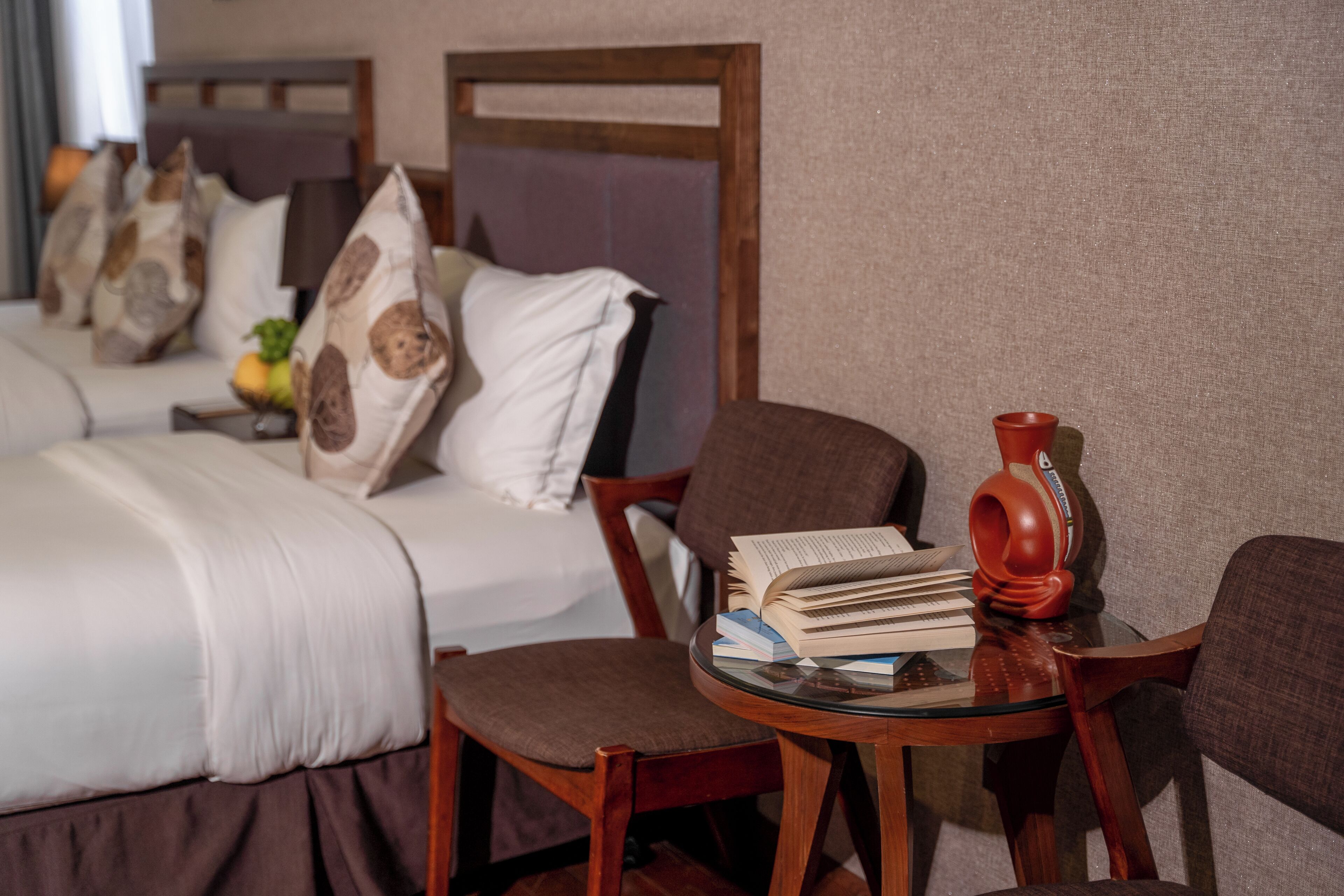 Ein Hotelzimmer mit einem Bett, einem Sessel, einem kleinen Tisch mit Büchern und einer Vase sowie einer Obstschale auf dem Nachttisch.