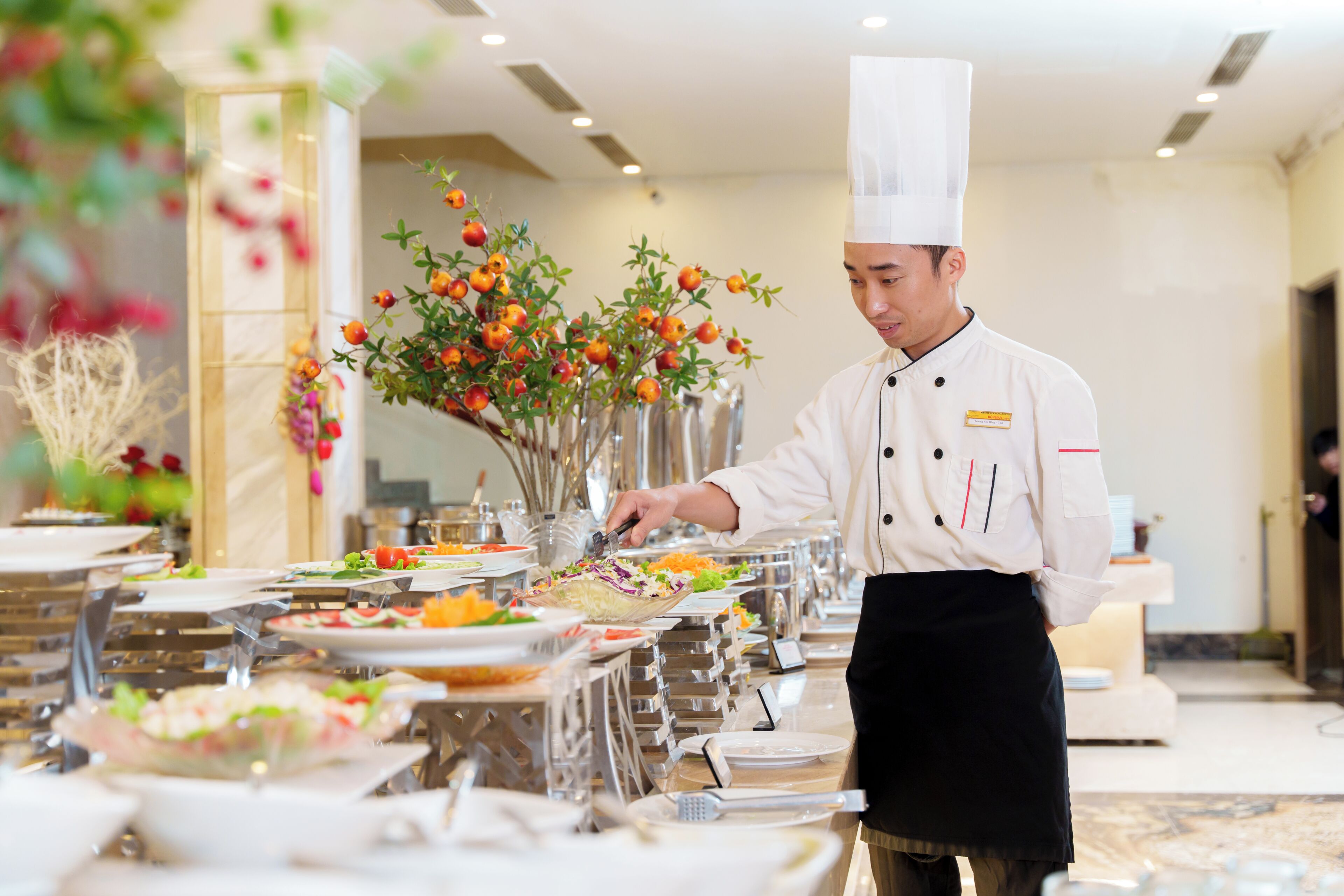A chef preparing food in a well-lit, spacious dining area with multiple tables and a decorative plant arrangement.