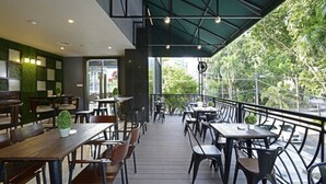 Un cafè a l'exterior amb taules i cadires de fusta, una paret verda i vista a arbres.