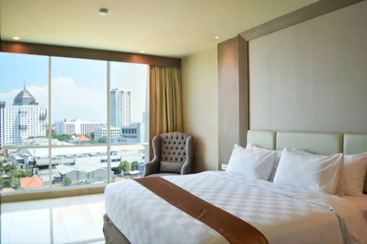 Ein Hotelzimmer mit einem großen Bett, einem Sessel und Blick auf eine Stadtlandschaft durch ein großes Fenster.