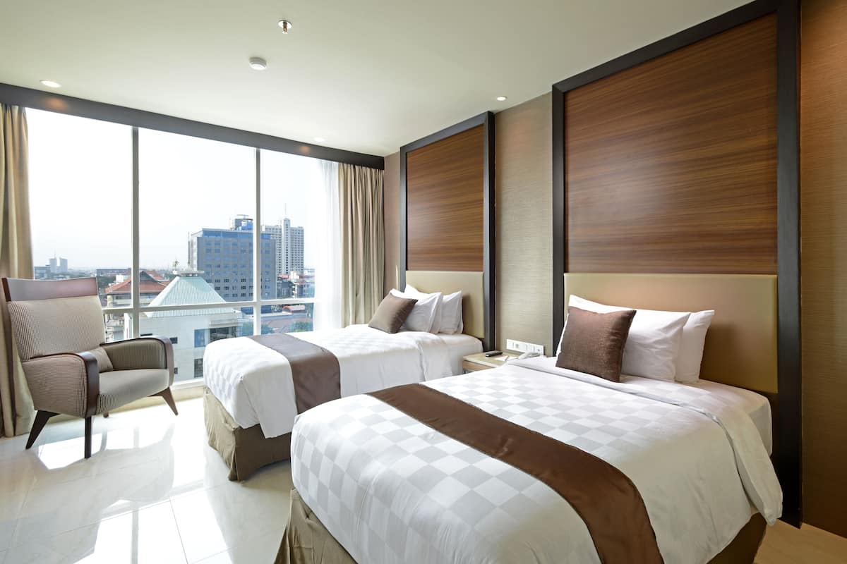 Ein Hotelzimmer mit zwei Betten, einem Sessel und einem großen Fenster mit Blick auf die Stadt.