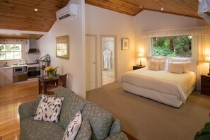 A bedroom with a large bed, a sitting area with a sofa, and a kitchen area visible through an open door. - Myers Creek Cascades Luxury Cottages (Healesville)