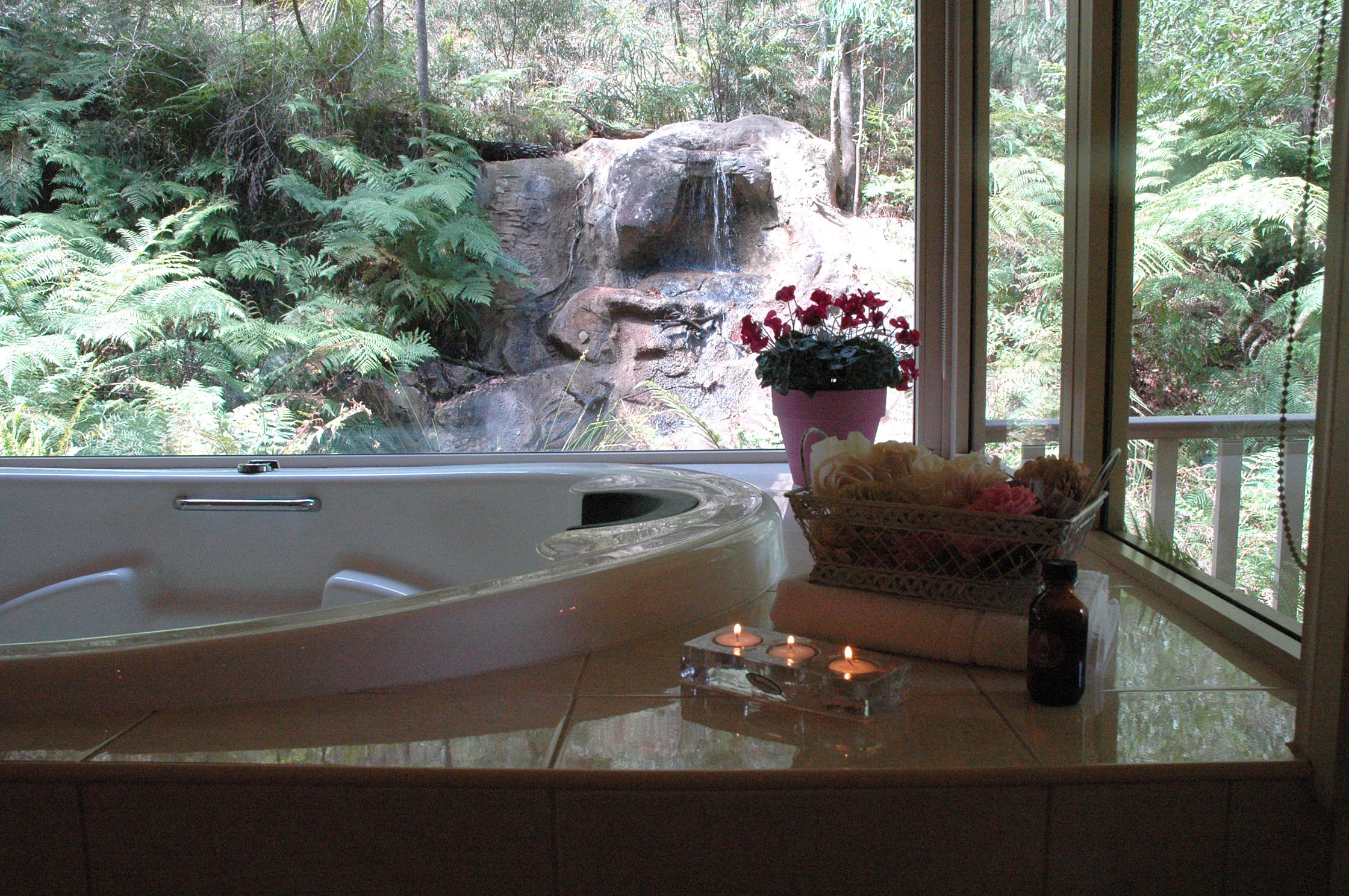 Une salle de bain avec une baignoire, une fenêtre donnant sur une cascade et la verdure, une plante en pot et des bougies posées sur le comptoir.