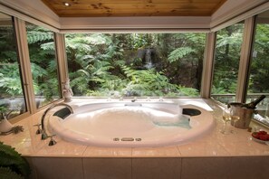 A large bathtub with a view of a forest through large windows. - Myers Creek Cascades Luxury Cottages (Healesville)