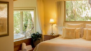 A hotel room with a large bed, a bedside table with a lamp, a window with curtains, a sink, and a towel on the floor. - Myers Creek Cascades Luxury Cottages (Healesville)