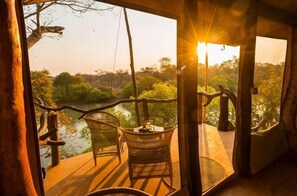 A wooden deck with wicker chairs overlooking a river and forest. - Mukambi Fig Tree Bush Camp – All Inclusive (Kafue National Park)